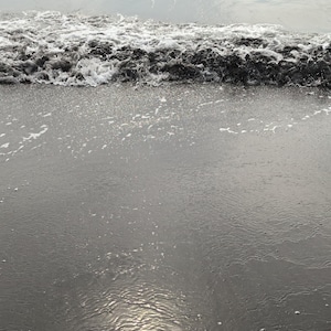 Könnte beinhalten: Graustufenbild eines Strandes. Eine Welle bricht am Ufer, mit weißem Schaum, der sich vom dunkelgrauen Wasser abhebt. Der nasse Sand reflektiert das Sonnenlicht und erzeugt einen schimmernden Effekt.