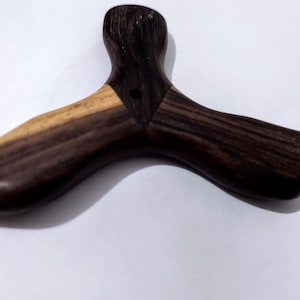 May include: A three-pronged wooden object with a smooth, polished finish. The piece features a dark brown wood grain throughout, with a contrasting lighter tan wood section on one of the three arms. A small, circular hole is centered at the junction.