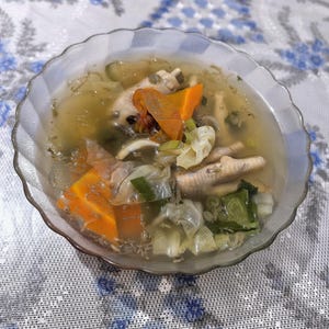 May include: A clear glass bowl with a scalloped rim containing a savoury soup. The broth features visible chicken feet, bright orange carrot chunks, chopped green leafy vegetables, and translucent cabbage pieces in a light, seasoned liquid base.