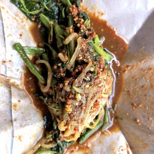 Puede incluir: Una porción de espinaca de agua salteada, conocida como kangkong, cubierta con una generosa cantidad de hojuelas de chile rojo triturado y especias tostadas, bañada en una salsa marrón oscura y sabrosa sobre papel pergamino blanco doblado.