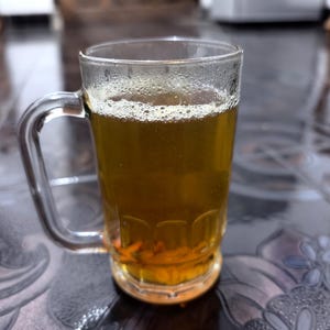 Könnte beinhalten: Ein klarer Glasbecher mit einem stabilen Henkel, gefüllt mit einer durchscheinenden goldbraunen Flüssigkeit. Die Oberfläche des Getränks zeigt eine dünne Schicht kleiner Blasen, und das Glas hat ein schlichtes Design mit einem dicken Boden.