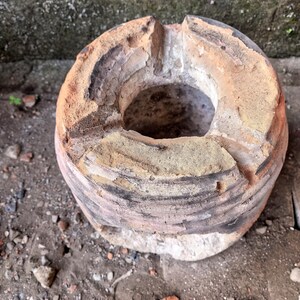 May include: A cylindrical clay stove with a weathered, textured surface featuring shades of tan, brown, and charcoal gray. The top rim has three distinct notches and a central circular opening, showing signs of heavy use and heat exposure.