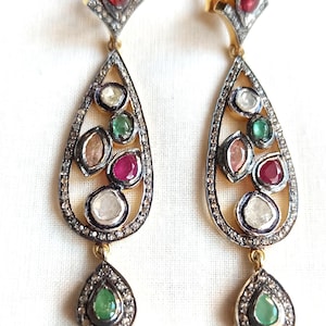 May include: A pair of teardrop-shaped earrings with a gold and silver setting. Each earring features a cluster of colorful gemstones, including red, green, and clear stones, surrounded by small, sparkling accents. The earrings have a dangling emerald-colored stone at the bottom.