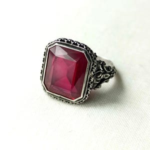 Op de afbeelding: Een zilveren ring met een grote, rechthoekige, robijnrode edelsteen. De ring heeft een sierlijke, filigraan-achtige zetting met ingewikkelde details rond de edelsteen en op de band. De edelsteen heeft een levendige, dieprode kleur.