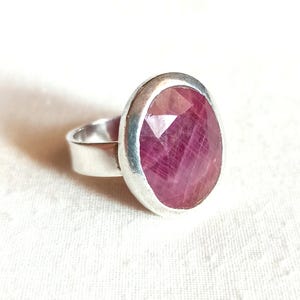Op de afbeelding: Een zilveren ring met een grote, ovaal geslepen robijn. De robijn heeft een facetgeslepen oppervlak, met roze en rode tinten. De band van de ring is van eenvoudig, gepolijst zilver. De edelsteen is gezet in een bezelzetting.