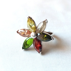 May include: A silver ring featuring a flower design with colorful marquise-cut gemstones. The petals are in shades of yellow, orange, green, and red, with a small silver bead at the center. The ring is set against a white background.