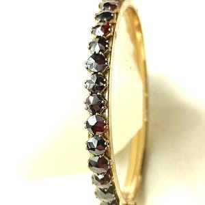 Pode incluir: Uma pulseira de bracelete dourada com uma fileira de pedras preciosas vermelho escuro. As pedras preciosas, de formato oval, são engastadas juntas, criando uma linha contínua. A pulseira tem um design simples e elegante.