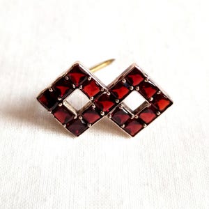 May include: A vintage brooch featuring two interlocking diamond-shaped sections. Each section is filled with small, square, deep red gemstones. The brooch has a gold-colored pin on the back and is set against a white background.