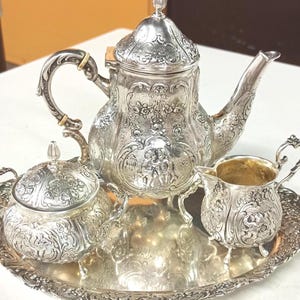 Puede incluir: Un juego de té de tono plateado que consta de una tetera con tapa, un azucarero con tapa, una jarra de leche y una bandeja ovalada. Cada pieza presenta intrincados patrones florales en relieve y escenas figurativas de querubines.