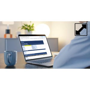 May include: A laptop displaying a spreadsheet with a blue and white color scheme. A blue mug sits to the left of the laptop. The laptop is on a wooden desk. A person in a blue shirt is visible in the background.