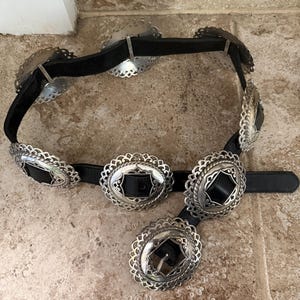 May include: A black belt with silver-colored, ornate oval and round buckles. The buckles have a decorative, scalloped edge and a black leather center. The belt is made of black leather and is displayed on a neutral-toned surface.
