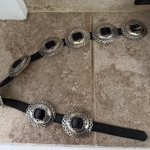 May include: A black belt with silver-colored oval-shaped buckles. The buckles have a decorative, openwork design around a black center. The belt is lying on a light brown tile surface.