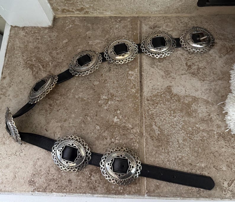 May include: A black belt with silver-colored oval-shaped buckles. The buckles have a decorative, openwork design around a black center. The belt is lying on a light brown tile surface.
