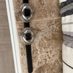 May include: A black belt with two ornate silver buckles. Each buckle has a black center and a decorative, scalloped edge. The belt is displayed against a neutral-toned background.