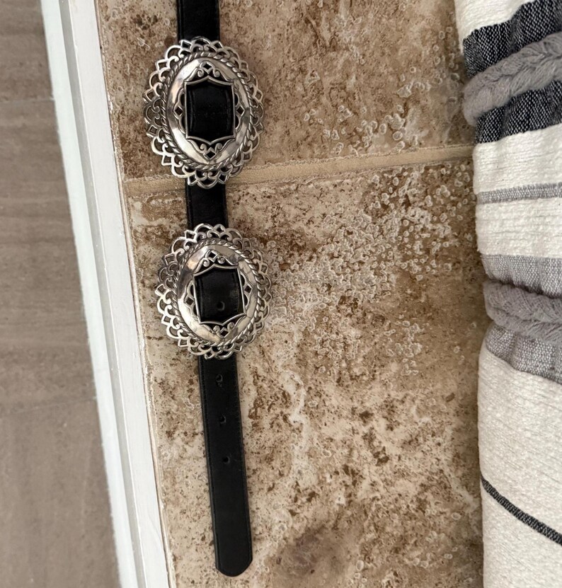 May include: A black belt with two ornate silver buckles. Each buckle has a black center and a decorative, scalloped edge. The belt is displayed against a neutral-toned background.