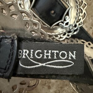 May include: Close-up of a black leather belt with a silver-toned buckle. The buckle has a central black square and an ornate, openwork design. The brand name "BRIGHTON" is visible on the belt.