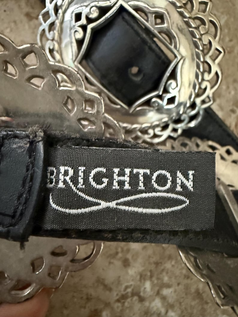 May include: Close-up of a black leather belt with a silver-toned buckle. The buckle has a central black square and an ornate, openwork design. The brand name "BRIGHTON" is visible on the belt.