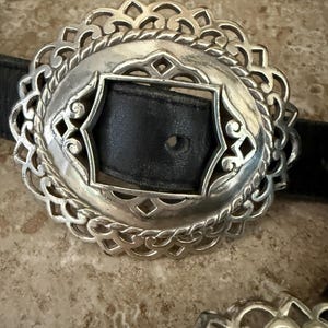 May include: Silver-toned oval belt buckle with an intricate filigree design. The buckle features a black leather strap insert. The decorative metalwork has a detailed, openwork pattern, creating a vintage aesthetic.