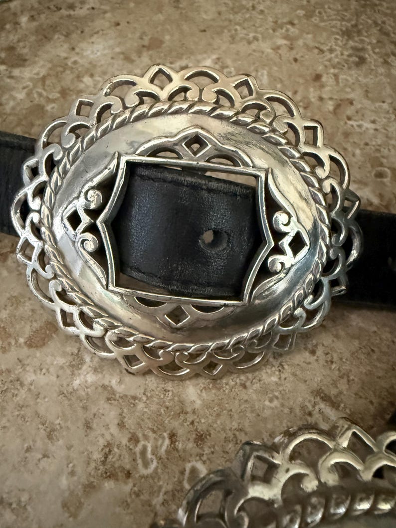 May include: Silver-toned oval belt buckle with an intricate filigree design. The buckle features a black leather strap insert. The decorative metalwork has a detailed, openwork pattern, creating a vintage aesthetic.