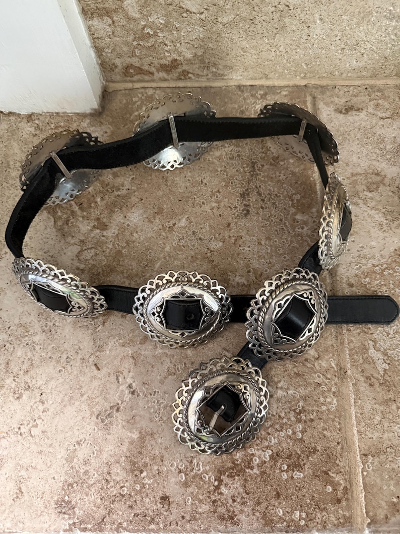 May include: A black belt with silver-colored, ornate oval and round buckles. The buckles have a decorative, scalloped edge and a black leather center. The belt is made of black leather and is displayed on a neutral-toned surface.