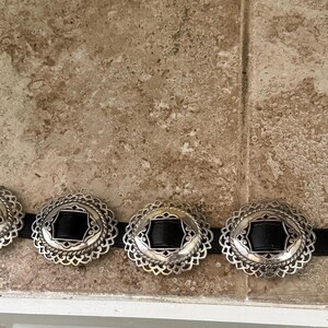 May include: A silver-toned belt with five decorative buckles. Each buckle has a black center and an ornate, scalloped edge. The belt is on a black strap. The buckles are arranged in a row.