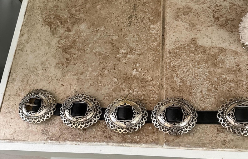 May include: A silver-toned belt with five decorative buckles. Each buckle has a black center and an ornate, scalloped edge. The belt is on a black strap. The buckles are arranged in a row.