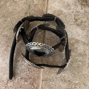 May include: A black belt with silver-colored decorative elements. The belt features multiple silver-toned, scalloped-edge medallions and a central, larger medallion. The belt's design suggests a Western or bohemian style.