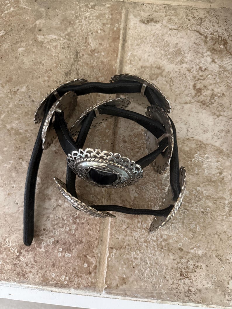 May include: A black belt with silver-colored decorative elements. The belt features multiple silver-toned, scalloped-edge medallions and a central, larger medallion. The belt's design suggests a Western or bohemian style.