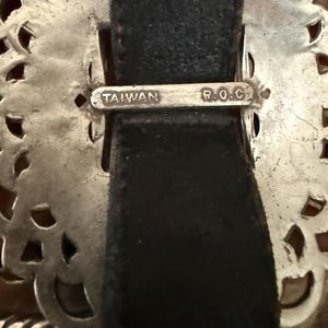May include: Close-up of a silver-toned belt buckle with an ornate, openwork design. The buckle is attached to a black leather belt. The text "TAIWAN R.O.C." is stamped on the buckle.