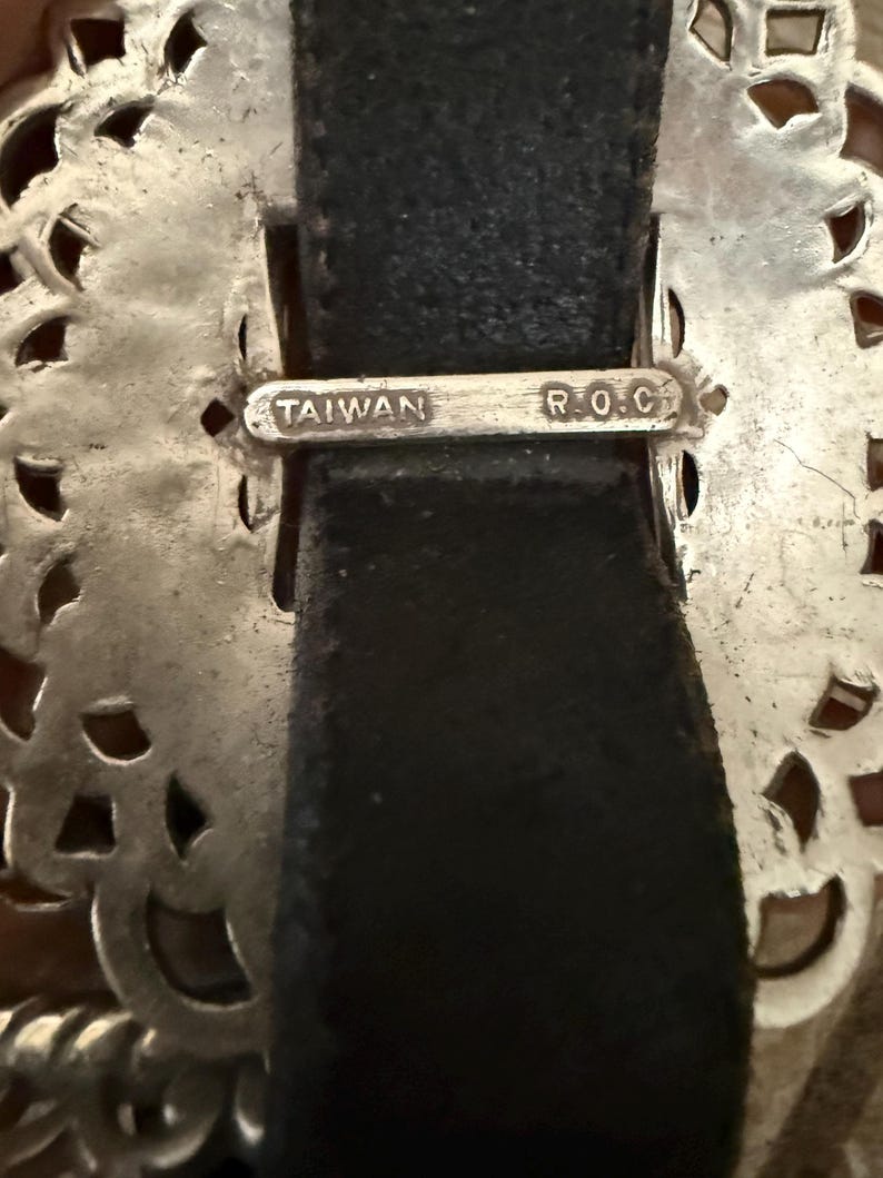 May include: Close-up of a silver-toned belt buckle with an ornate, openwork design. The buckle is attached to a black leather belt. The text "TAIWAN R.O.C." is stamped on the buckle.