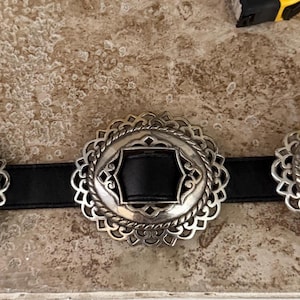 May include: A black leather belt with three silver-toned, ornate buckles. Each buckle has a black center and a decorative, scalloped edge. The belt is displayed on a textured, light brown surface.