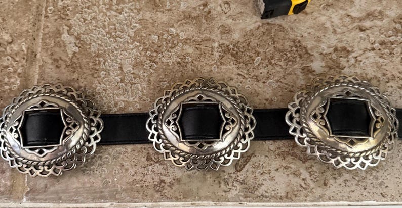 May include: A black leather belt with three silver-toned, ornate buckles. Each buckle has a black center and a decorative, scalloped edge. The belt is displayed on a textured, light brown surface.