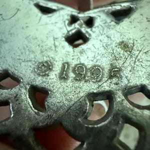 May include: Close-up of a silver-toned metal pendant with an openwork design. The pendant has a butterfly shape with cut-out details and the year "1995" stamped on it. The pendant is slightly tarnished.