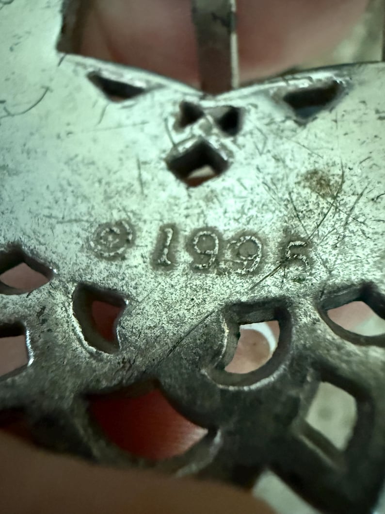 May include: Close-up of a silver-toned metal pendant with an openwork design. The pendant has a butterfly shape with cut-out details and the year "1995" stamped on it. The pendant is slightly tarnished.