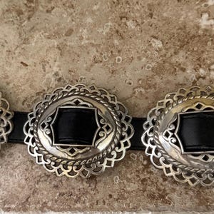 May include: A black leather belt with silver-colored decorative buckles. The buckles have a scalloped edge design with a central black leather insert. The belt buckle has a silver-colored metal clasp.