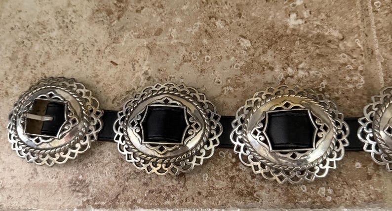 May include: A black leather belt with silver-colored decorative buckles. The buckles have a scalloped edge design with a central black leather insert. The belt buckle has a silver-colored metal clasp.