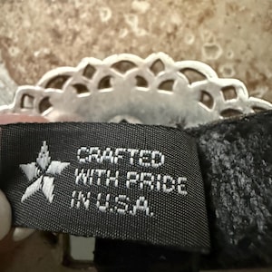 May include: Close-up of a black tag with white text that reads "CRAFTED WITH PRIDE IN U.S.A." A silver-colored, decorative, scalloped edge is visible above the tag. The background is a mottled, light brown and tan surface.