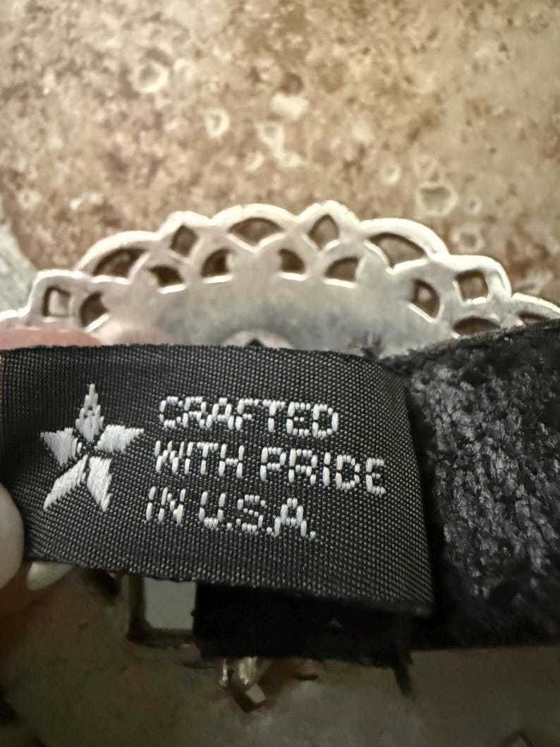 May include: Close-up of a black tag with white text that reads "CRAFTED WITH PRIDE IN U.S.A." A silver-colored, decorative, scalloped edge is visible above the tag. The background is a mottled, light brown and tan surface.
