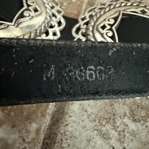 May include: Black leather belt with two ornate silver-colored buckles. The buckles have a filigree design. The belt has the stamped text "M 36603".