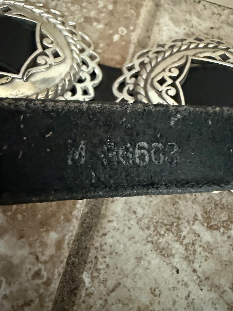 May include: Black leather belt with two ornate silver-colored buckles. The buckles have a filigree design. The belt has the stamped text "M 36603".