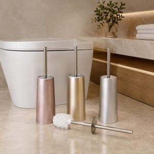 Op de afbeelding: Drie toiletborstelsets met metalen houders in roségoud, goud en zilver. Elke set bevat een lange, slanke roestvrijstalen steel en een bijpassende houder. Eén borstel ligt horizontaal voor de houders op de vloer.