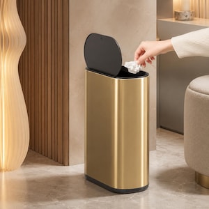 Könnte beinhalten: Ein hoher, ovaler Abfalleimer mit metallisch goldener Oberfläche und einem mattschwarzen Deckel. Der Behälter hat ein schlankes, aufrechtes Design mit flachem Boden und einem Klappdeckel, der sich nach oben öffnet und das dunkle Innere zeigt.