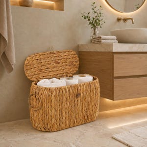 Op de afbeelding: Een ovale opbergmand gevlochten van natuurlijke lichtbruine zeegrasvezels. De mand heeft een gedeeltelijk geopend scharnierend deksel, waardoor twee witte rollen toiletpapier aan de binnenkant zichtbaar zijn. Aan de zijkant zit een ronde handgreep.