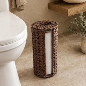 Op de afbeelding: Een cilindrische toiletrolhouder gemaakt van donkerbruin gevlochten zeegras. De verticale opbergeenheid is voorzien van een afneembaar rond deksel met een kleine handgreep en een open zijgleuf, met daarin drie gestapelde rollen wit toiletpapier.