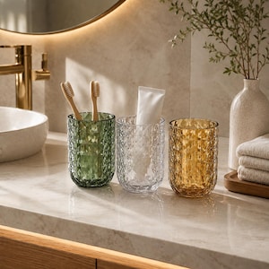 May include: Three cylindrical glass tumblers with a faceted diamond-cut texture. One tumbler is green and holds two wooden toothbrushes, one is clear and holds a white tube of toothpaste, and the third is amber-coloured and stands empty.
