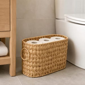Puede incluir: Una cesta de almacenamiento ovalada tejida con fibras naturales de color tostado claro, que cuenta con un asa circular resistente en el lateral. La cesta contiene tres rollos de papel higiénico blancos alineados, ofreciendo una solución funcional.