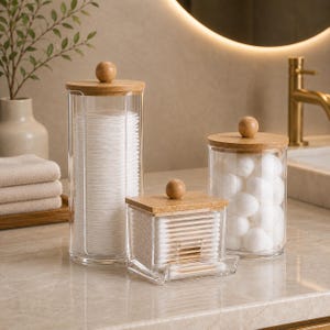 Puede incluir: Un juego de tres organizadores de baño de acrílico transparente con tapas de madera clara y pomos esféricos. La colección incluye un recipiente cilíndrico alto, una caja cuadrada y un tarro más corto para diversos artículos de aseo.