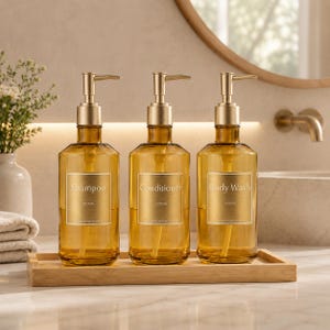 Könnte beinhalten: Drei bernsteinfarbene Glasflaschen mit Pumpmechanismus und goldfarbenen Metallspendern, jeweils mit einem rechteckigen Etikett mit der Aufschrift Shampoo, Conditioner oder Body Wash, auf einem rechteckigen Bambustablett.