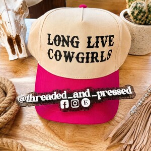 Op de afbeelding: Een beige en felroze baseballpet met de zwarte tekst "LONG LIVE COWGIRLS". De pet heeft een felroze klep en een roze knop aan de bovenkant. De afbeelding bevat ook een decoratief bord met social media handles.