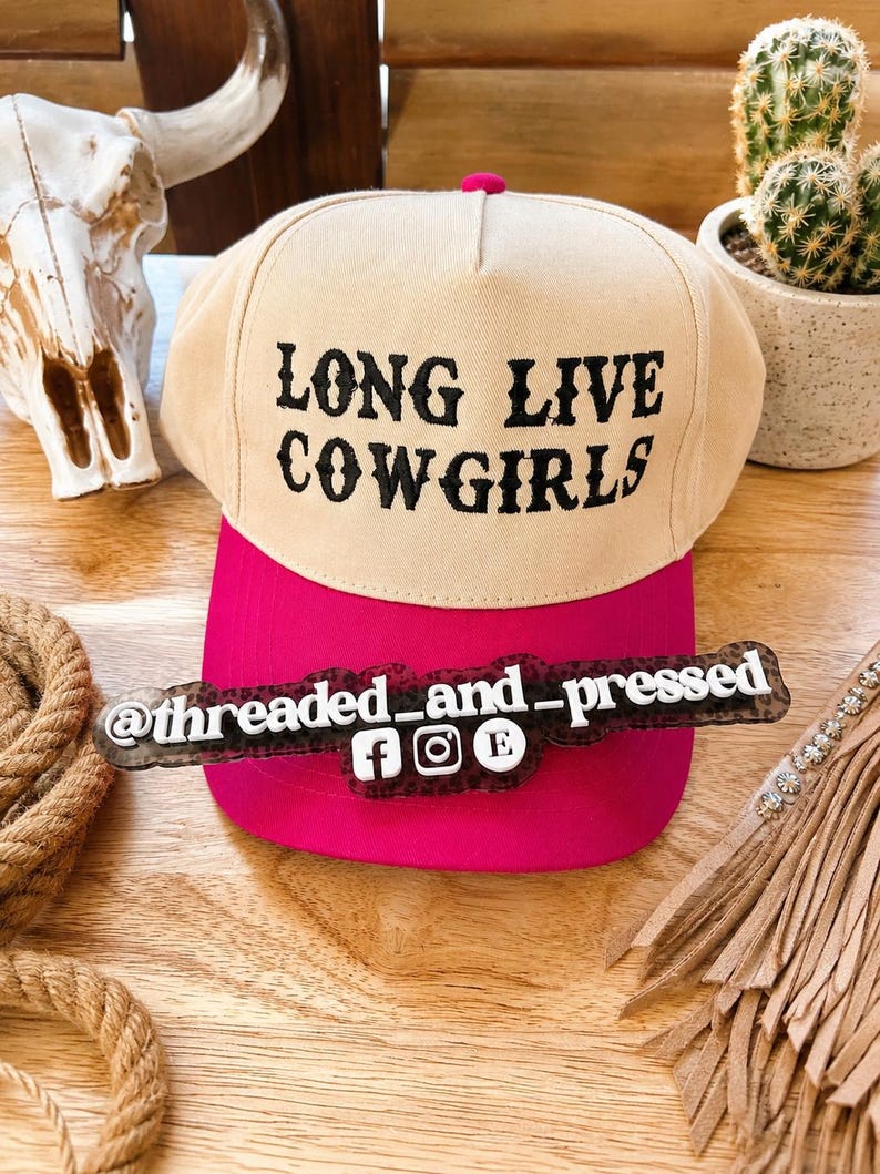 Op de afbeelding: Een beige en felroze baseballpet met de zwarte tekst "LONG LIVE COWGIRLS". De pet heeft een felroze klep en een roze knop aan de bovenkant. De afbeelding bevat ook een decoratief bord met social media handles.